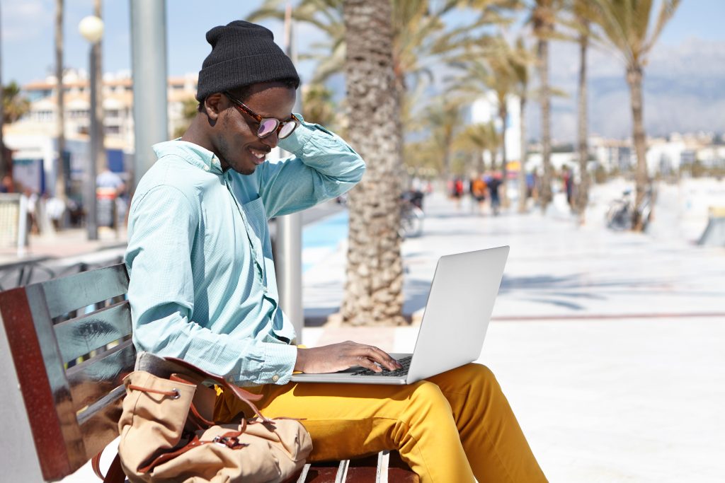 stylish-young-african-american-male-freelancer-wearing-hat-shades-using-laptop-remote-work-using-free-city-wireless-intern...